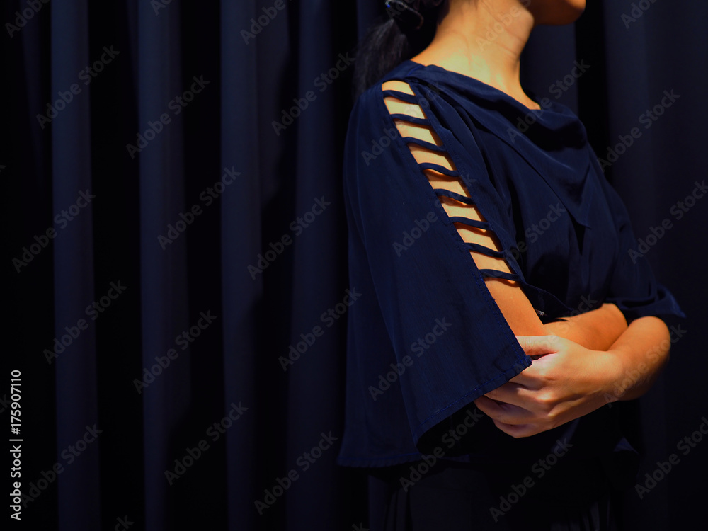 Woman wearing blue black shirt with shoulder hole details, with black curtain background, turn her body twist to the left as to escape, cross arms to embrace herself, showing disagree or quarrel