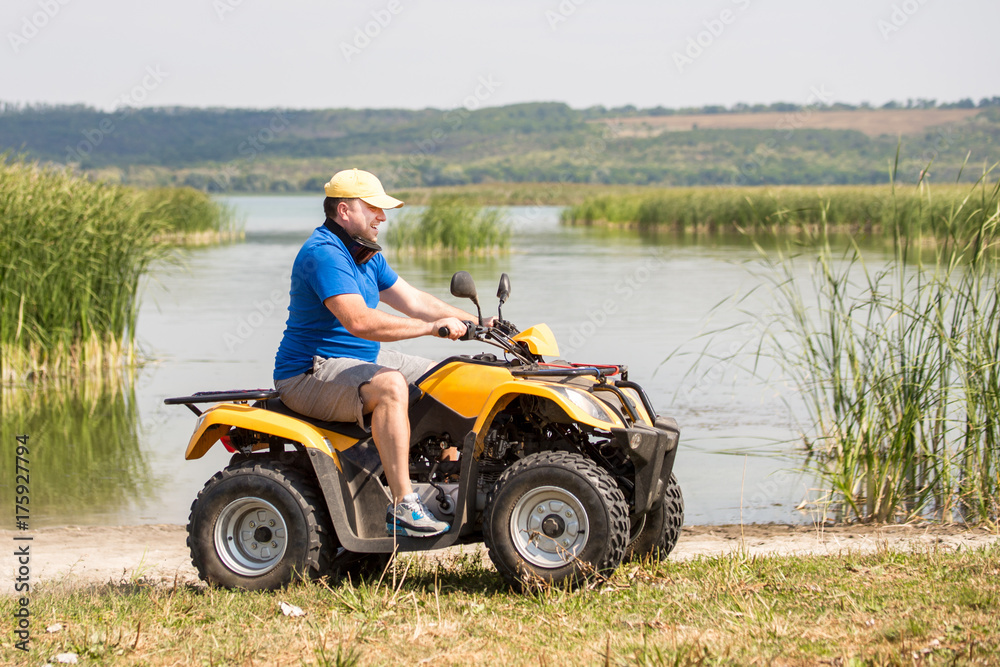 Caucasian man riding an ATV quad bike over rough terrain near lake or river shore with bulrush and reed. Adventure activity concept Caucasian man riding an ATV quad bike over rough terrain near lake or river shore with bulrush and reed. Adventure activity concept