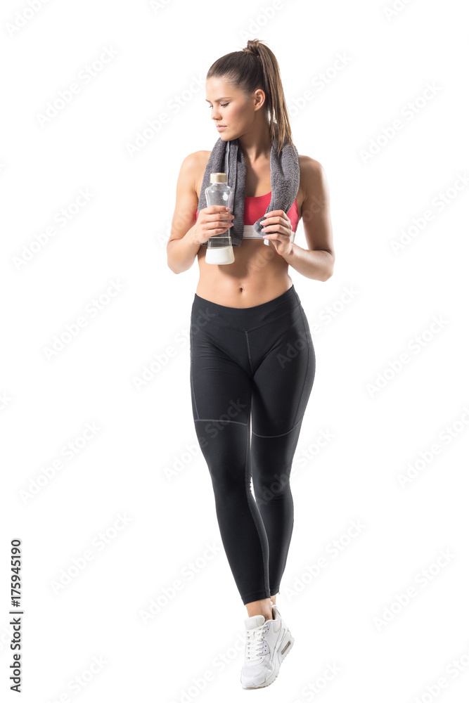 Confident healthy sportswoman in sportswear with towel and glass water bottle looking down. Full body length portrait isolated on white background. Confident healthy sportswoman in sportswear with towel and glass water bottle looking down. Full body length portrait isolated on white background.