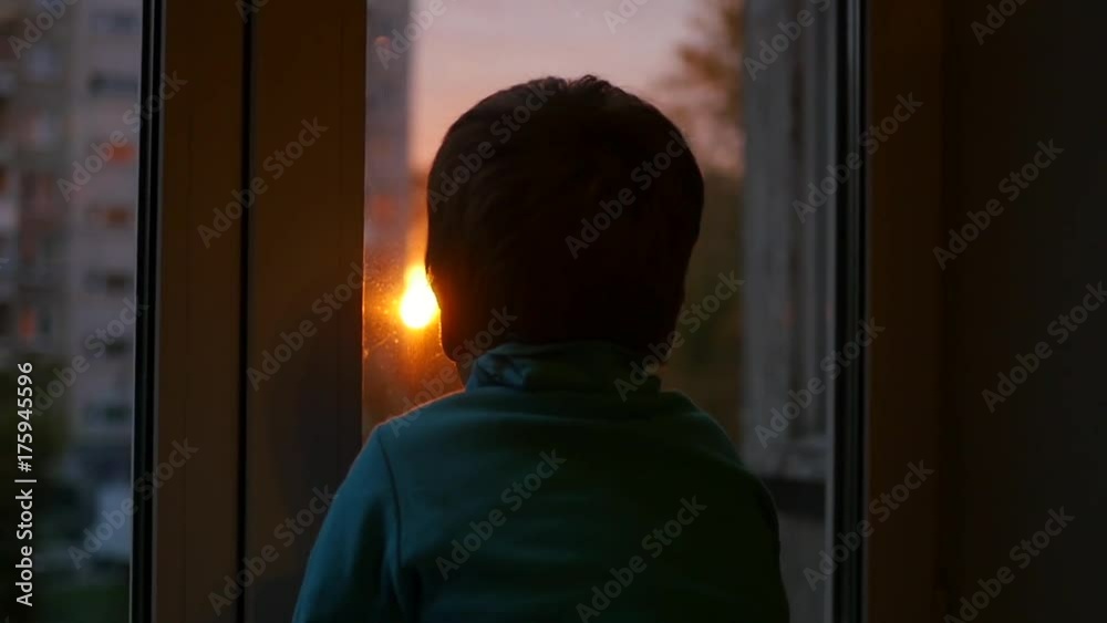 a child stands on the windowsill and looking out the window at the sun. Sunset a child stands on the windowsill and looking out the window at the sun. Sunset
