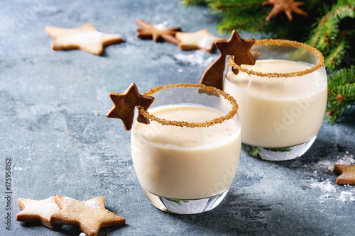 Eggnog Christmas milk cocktail with cinnamon, served in two glasses with shortbread star shape sugar cookies different size, decor toys, fir branch over blue texture background.