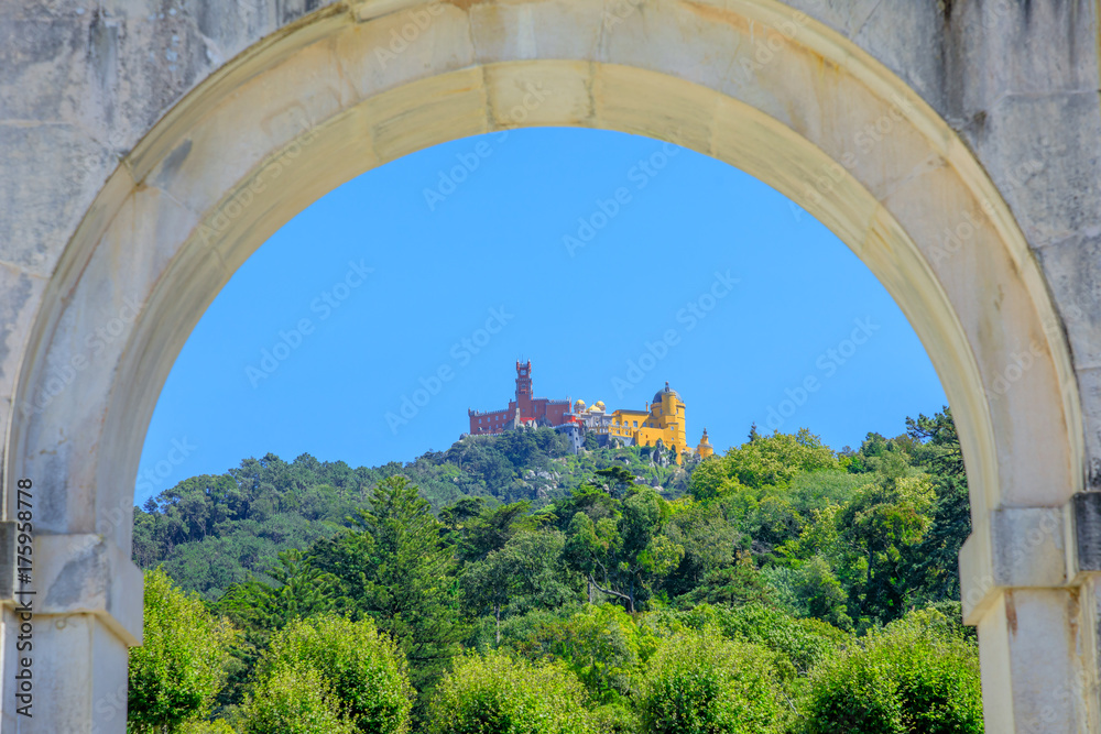 Fototapeta premium Close up of Pena National Palace in the blue sky surrounded by green hills, seen from the arch of Seteais Palace. Sintra historic center, Lisbon District, Portugal.