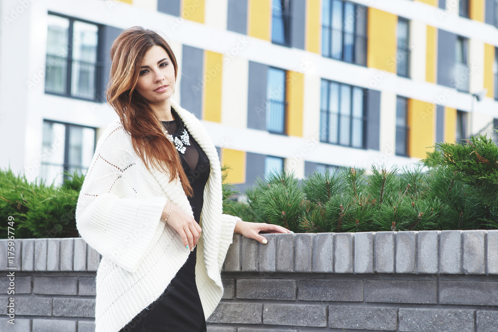 Beautiful young fashionable woman in a black short dress and white cardigan posing in the city. Fashion photo