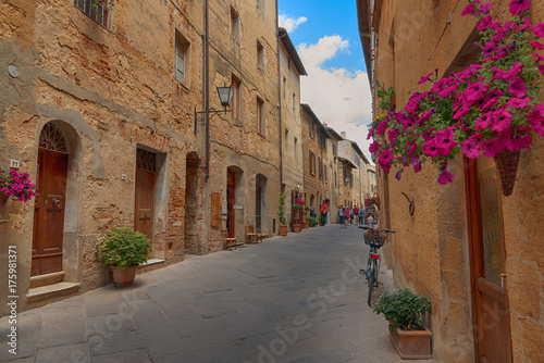 Fototapeta Naklejka Na Ścianę i Meble -  Beautiful narrow street with sunlight and flowers in the small magical and old village of Pienza, Val D'Orcia Tuscany, Italy.
