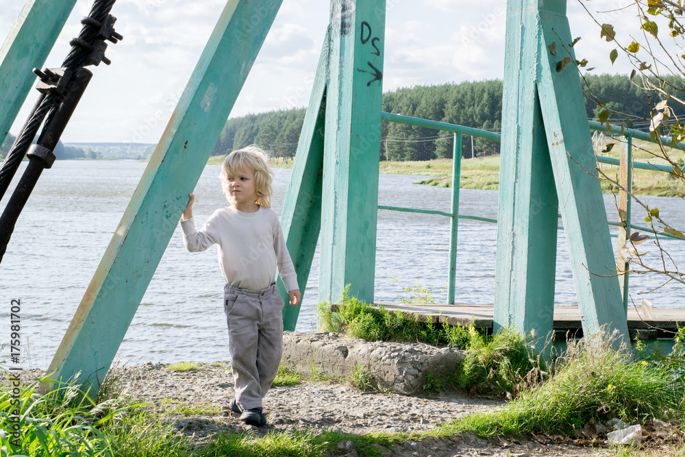 A happy blond boy staying at an Old wooden pendant bridge - summertime activity at a country village, the Urals, Russiaf, Kuryi