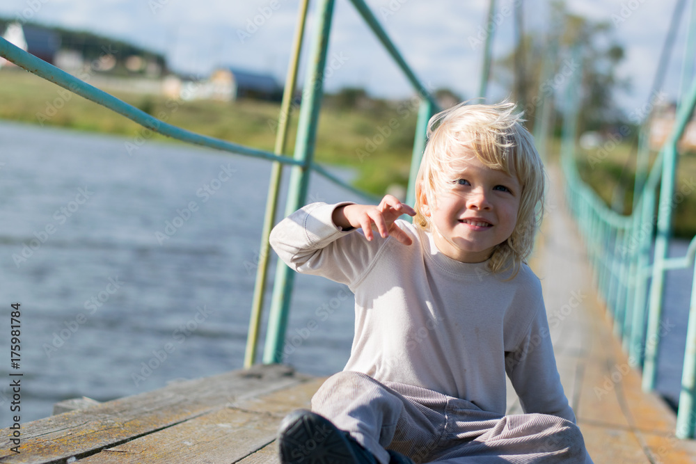 A happy blond boy sitting at an Old wooden pendant bridge - summertime activity at a country village, the Urals, Russiaf, Kuryi