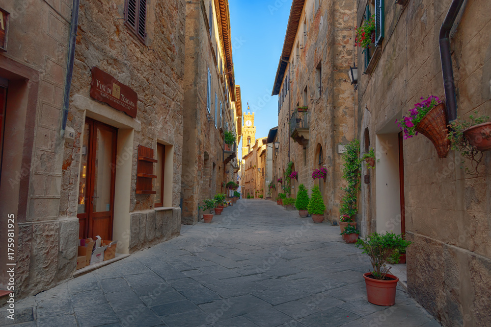 Beautiful narrow street with sunlight and flowers in the small magical and old village of Pienza, Val D'Orcia Tuscany, Italy.