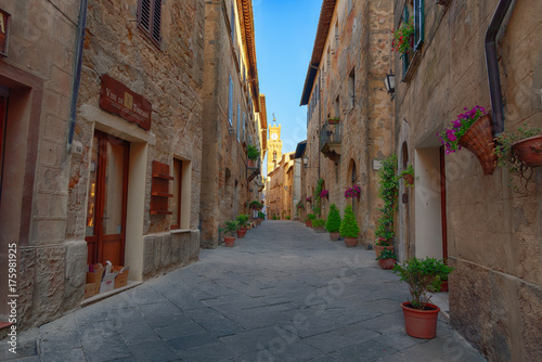 Beautiful narrow street with sunlight and flowers in the small magical and old village of Pienza, Val D'Orcia Tuscany, Italy. © djevelekova