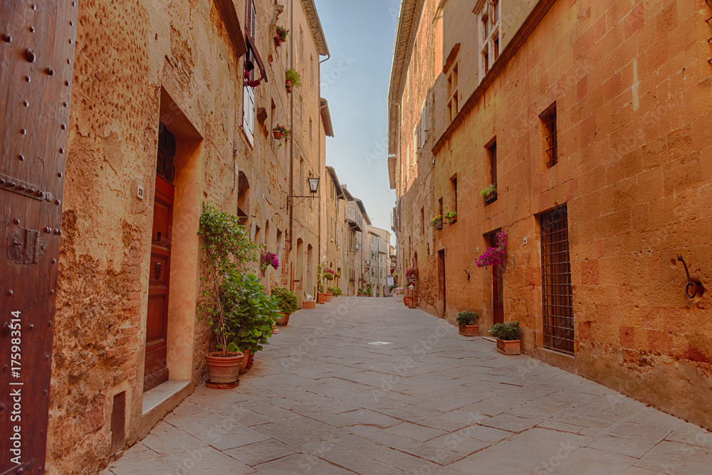 Obraz premium Beautiful narrow street with sunlight and flowers in the small magical and old village of Pienza, Val D'Orcia Tuscany, Italy.