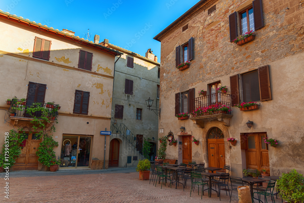 Obraz premium Beautiful narrow street with sunlight and flowers in the small magical and old village of Pienza, Val D'Orcia Tuscany, Italy.