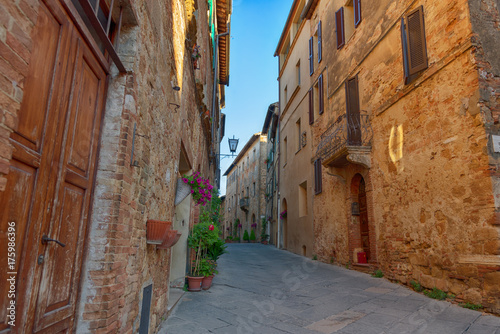 Fototapeta Naklejka Na Ścianę i Meble -  Beautiful narrow street with sunlight and flowers in the small magical and old village of Pienza, Val D'Orcia Tuscany, Italy.