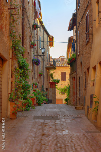 Fototapeta Naklejka Na Ścianę i Meble -  Beautiful narrow street with sunlight and flowers in the small magical and old village of Pienza, Val D'Orcia Tuscany, Italy.