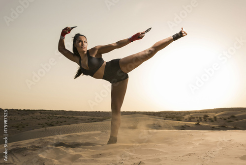 A Beautiful Asian-Black, Mixed Race martial artist female performs kicks, punches and stick fighting with creative leaps in the desert at sunrise or sunset wearing black short tights and black sports 