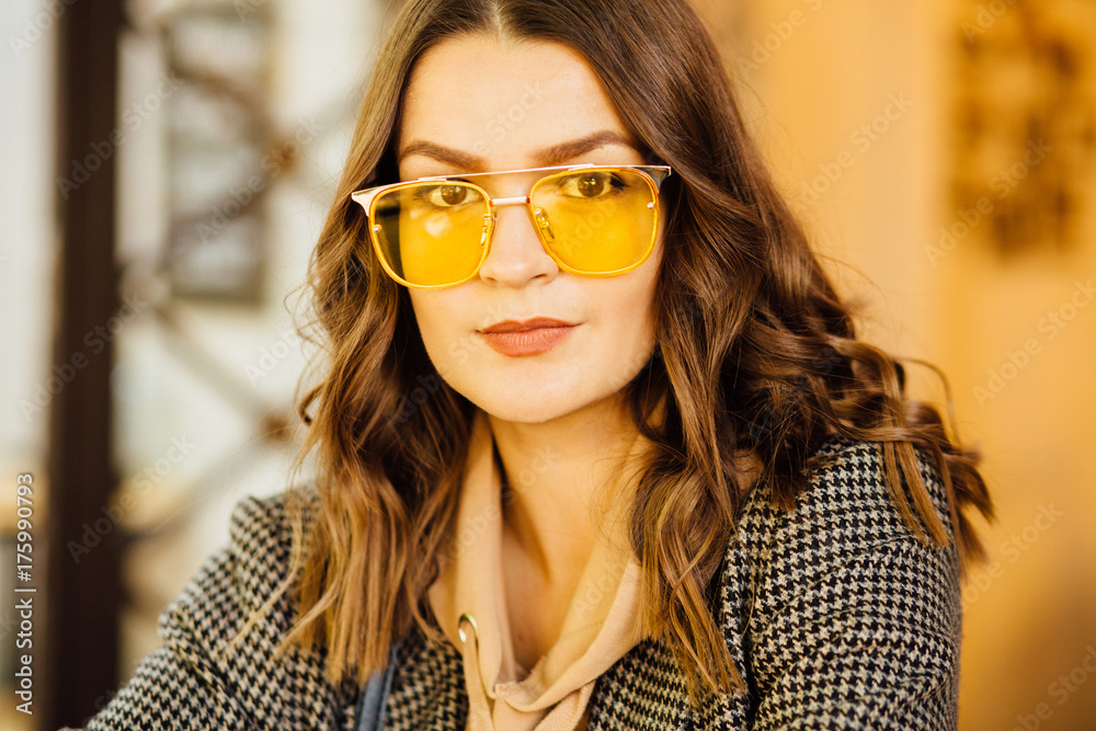 Close up portrait of fashionable hipster woman in yellow sunglasses and in casual jacket looking at camera outdoor in blurred background of street cafe. Business woman, lifestyle and beauty concept. Close up portrait of fashionable hipster woman in yellow sunglasses and in casual jacket looking at camera outdoor in blurred background of street cafe. Business woman, lifestyle and beauty concept.