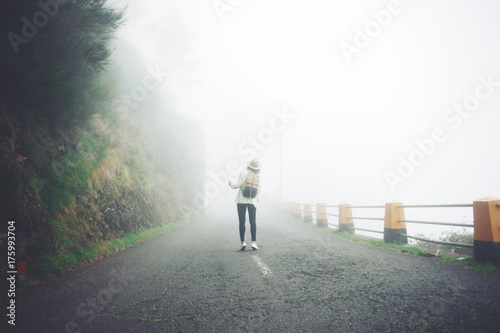 Young travelling woman wearing backpack and hat standing on the road in fog, searching path and using map.