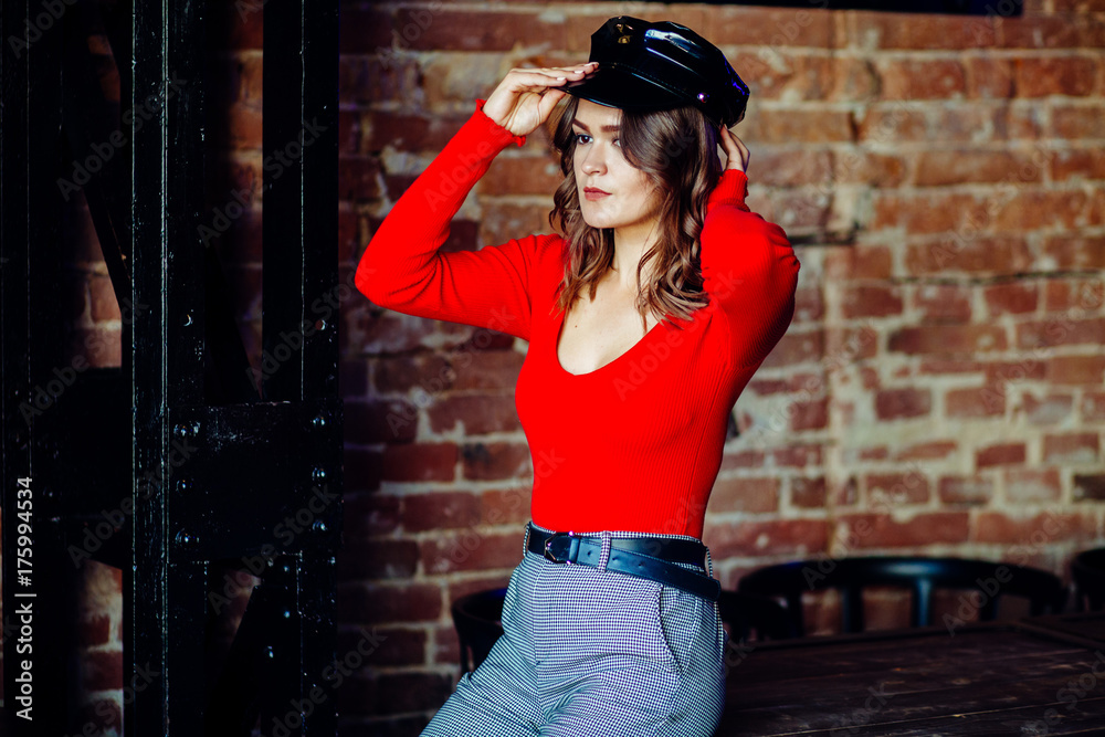 Obraz premium Provocated woman in red shirt and black peaked caps posing near in dark loft pub or night club interior.