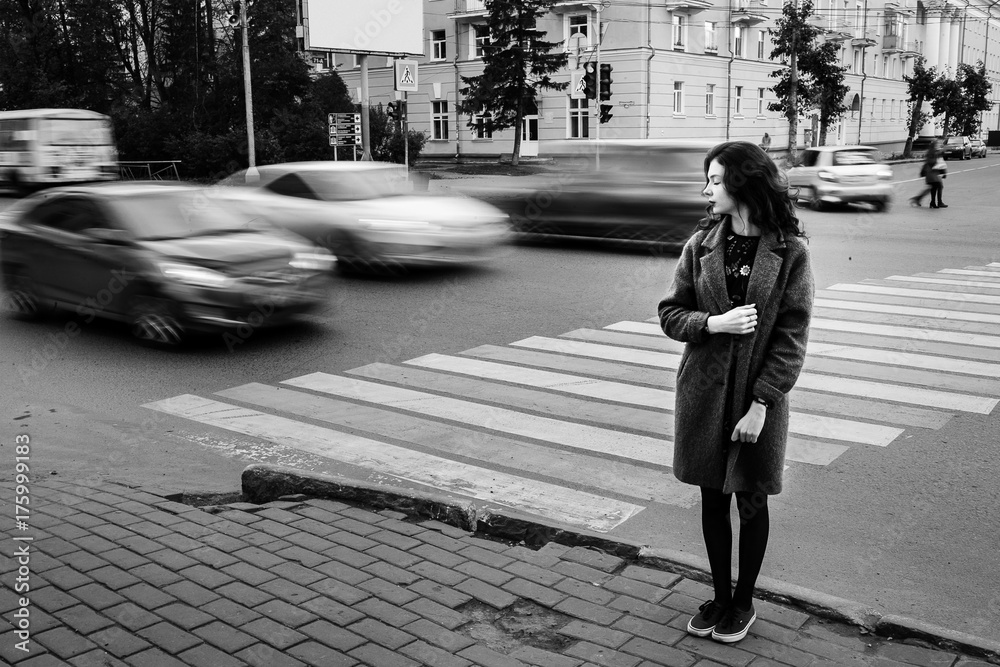Obraz premium One teenage girl blue coat standing at the traffic light on city street on a cloudly autumn day with vehicles passing by in motion blur.
