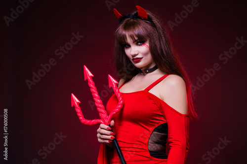 A beautiful, sexy woman in a devil costume, a demon with horns and pitchforks, a trident, in a red dress. American, dress for a costume party Halloween. Choker on the neck. Bright makeup
