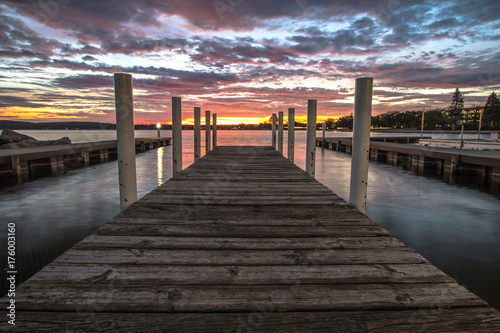 Wooden Dock On Sunrise Lake. Summer sunrise over the waters of Grand Traverse Bay in Traverse City, Michigan. Shot with long wooden dock in foreground sunrise over water at horizon.