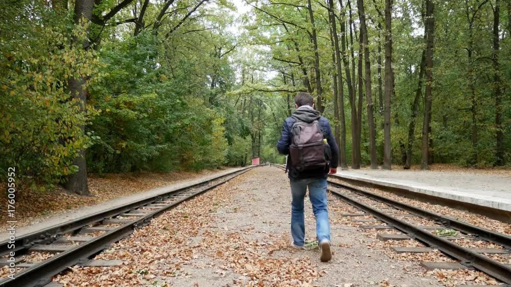Young man walks along the platform between the railway rails. A railroad station in the forest in the autumn. Student traveler with a black backpack and in a blue jacket walks away from the camera Young man walks along the platform between the railway rails. A railroad station in the forest in the autumn. Student traveler with a black backpack and in a blue jacket walks away from the camera
