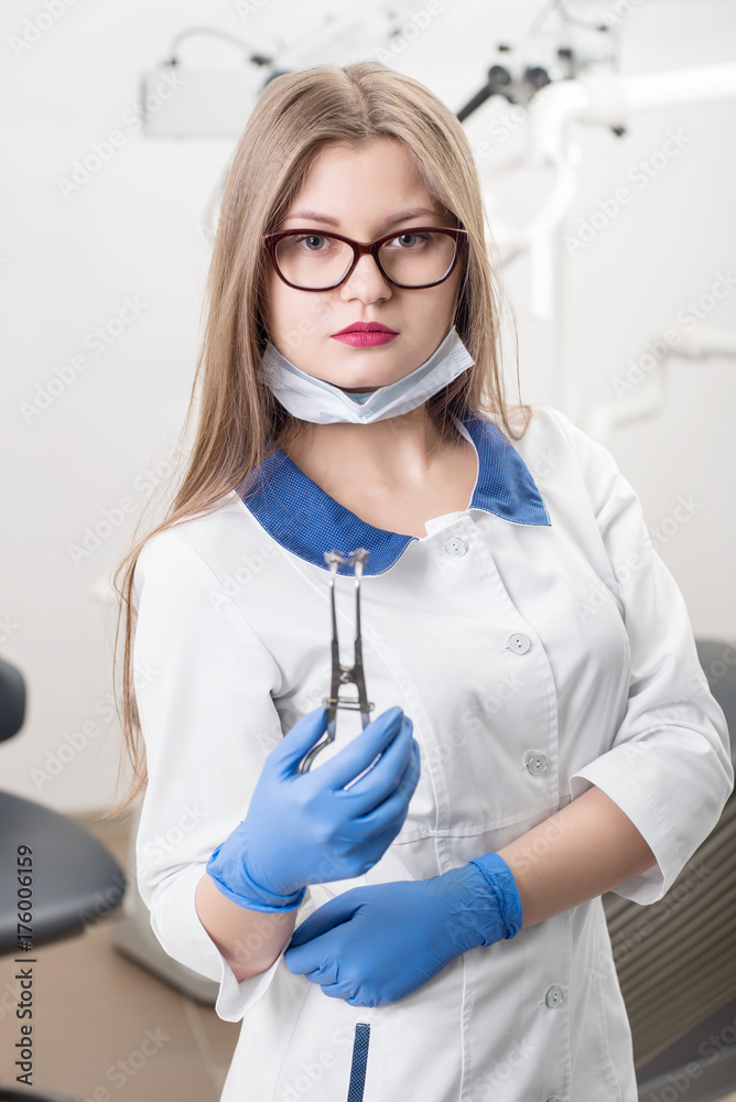 Portrait of young attractive woman dentist holding dental device at the modern dental office. Doctor wearing glasses, white uniform, blue gloves and looking to the camera. Dentistry