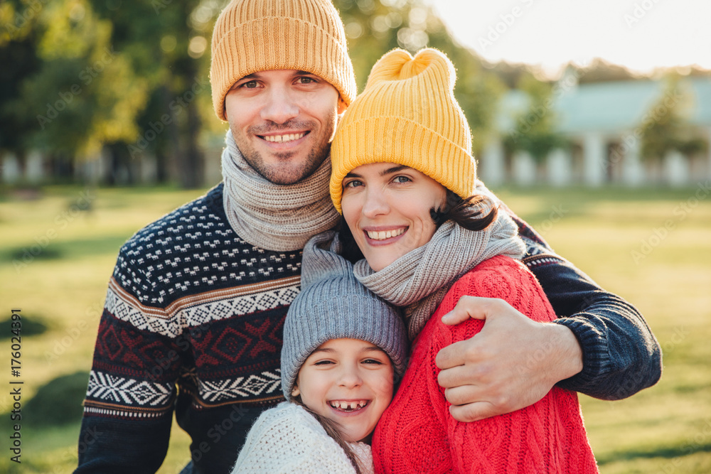 Obraz premium Adorable little child stand close to her affectionate parents, enjoy spending time together, embrace each other, smile happily, look directly into camera. Pleasant unforgettable moments with family