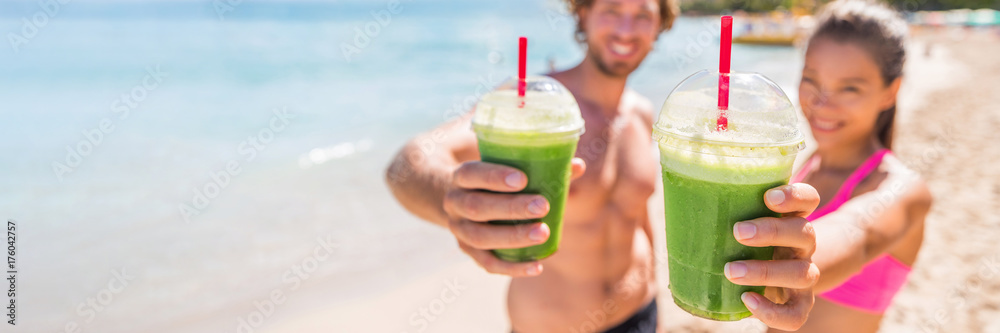 Fototapeta premium Fitness couple drinking green smoothie at beach banner panoramic. Man and woman holding vegetable smoothies after running sport fitness training. Healthy clean eating lifestyle concept.