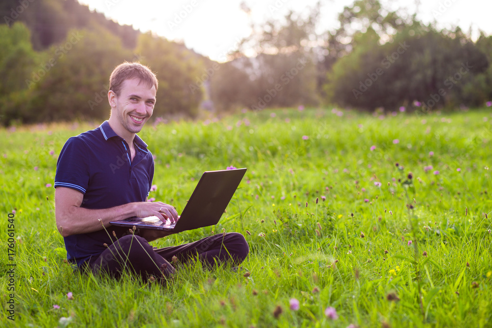 Young man sitting on green grass at sunset with laptop and working Young man sitting on green grass at sunset with laptop and working