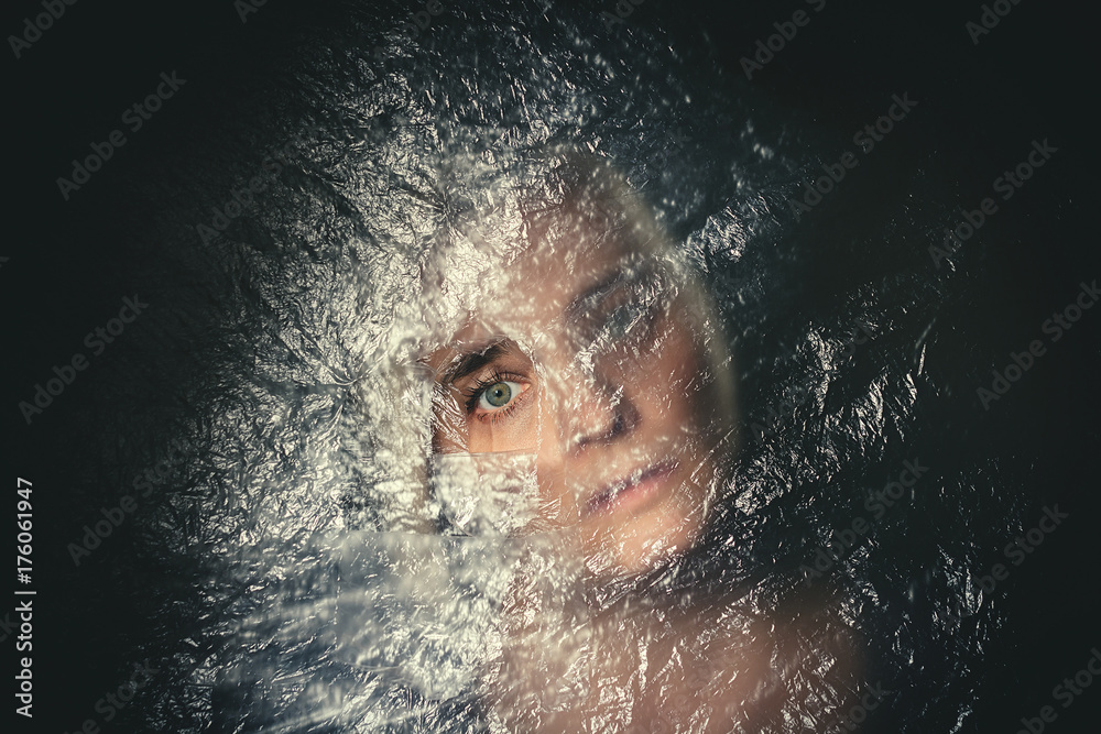 Woman with blank, terrified expression peeking through hole in plastic curtain, scared in dark