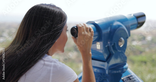 Anonymous brunette in casual outfit standing on viewpoint above city and exploring landscape with spyglass in sunlight.