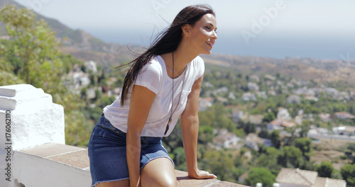 Pretty brunette in casual outfit looking excited while posing on viewpoint and exploring coastal city from height in summertime.