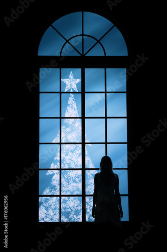 Woman watching snowy christmas tree through big window. Greeting card.