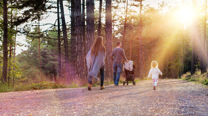 Happy young family taking a walk in a park, back view. Family holding hands walking together along forrest path with their daughter, father pushing the pram.