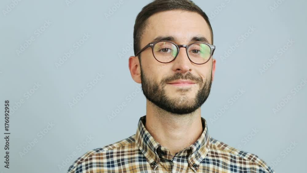 Close up shot of a young caucasian man in a casual shirt and glasses looking at the side and turning his face to the camera, smiling at camera against the grey background. Outdoor shot. Close up shot of a young caucasian man in a casual shirt and glasses looking at the side and turning his face to the camera, smiling at camera against the grey background. Outdoor shot.