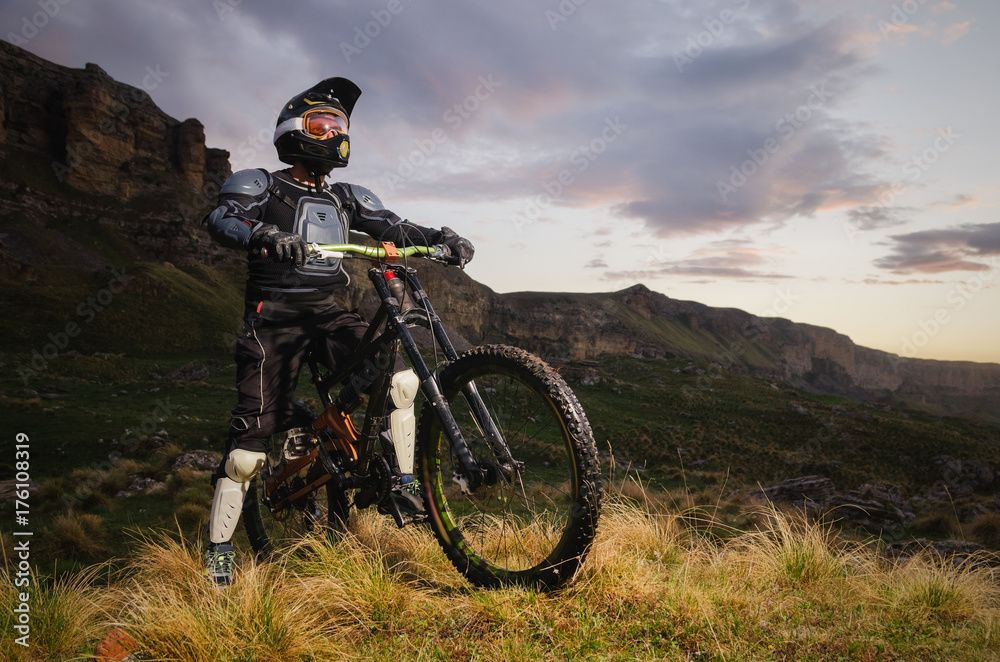 The rider in full protection on a mountain bike stands and looks at the sunset on the background of the rocks