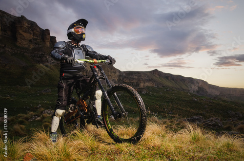 The rider in full protection on a mountain bike stands and looks at the sunset on the background of the rocks