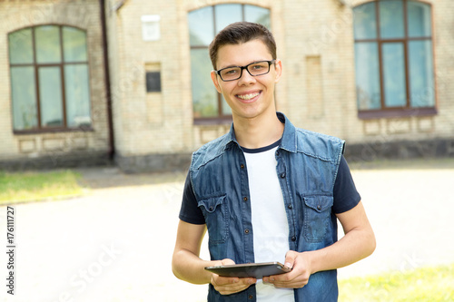 Male student wearing glasses is holding a digital tablet standing in front of the university. Happy young man. © A Stock Studio