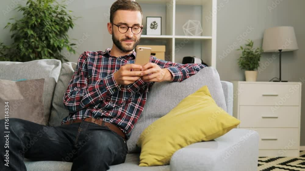 Young attractive man in glasses is sitting on the couch at home,holding the mobile phone and texting his friends via internet. Indoor. Young attractive man in glasses is sitting on the couch at home,holding the mobile phone and texting his friends via internet. Indoor.