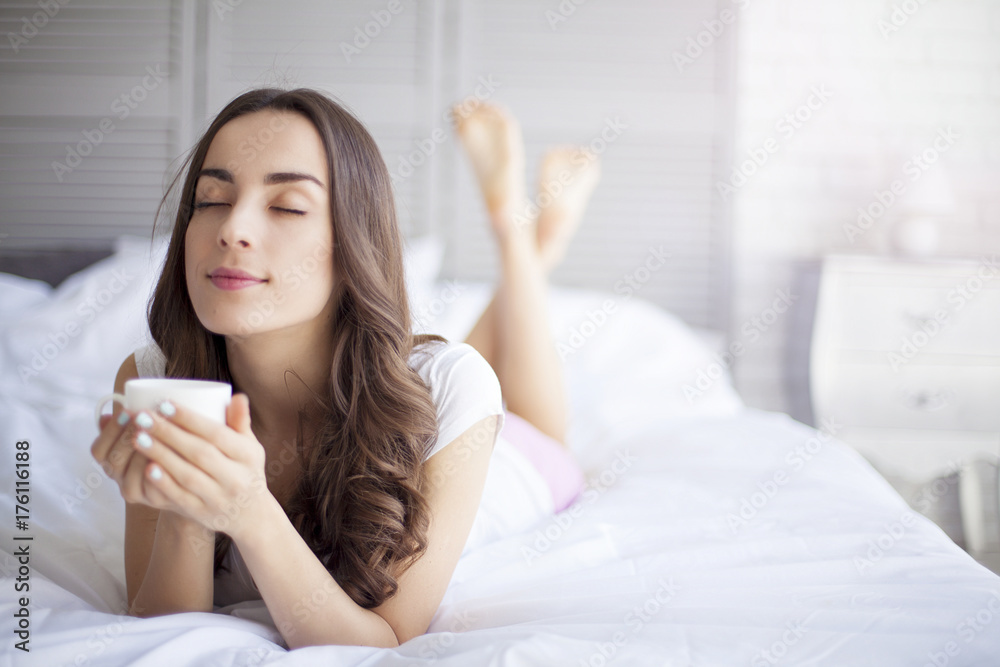 Attractive young smiling brunette woman lying in white bed and drinks a coffee in her bedroom. Attractive young smiling brunette woman lying in white bed and drinks a coffee in her bedroom.