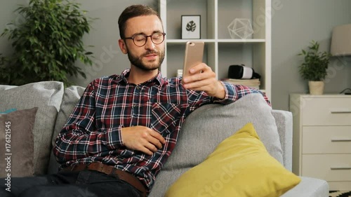 Handsome caucasian man in checked casula shirt and glasses is having a great time at home, sitting on the couch and videochatting with family at his free time. Indoor shot. photo