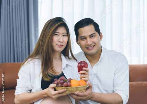 Handsome husband and his beautiful pregnant wife are relaxing together in the living room, husband give fruits to his wife