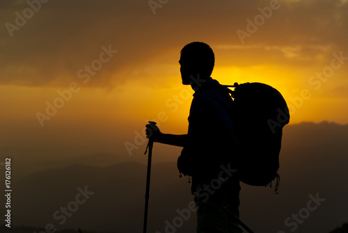 the silhouette of a man with a backpack, a trunk for the camera and trekking poles on the background of a sunset and a blurred mountain landscape