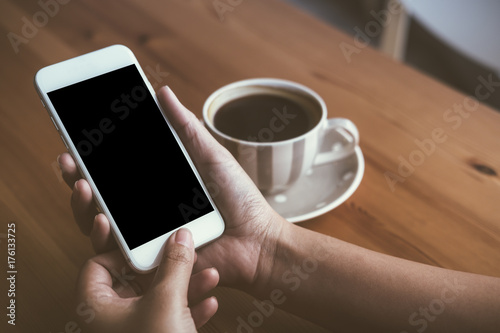Mockup image of woman's hands holding white mobile phone with blank black screen and white coffee cups in modern loft cafe
