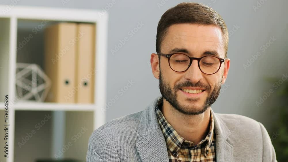 Close up face of an attractive caucasian man in spectacles, wearing a casual shirt and a grey shirt sitting at the office desk. Indoor shot. Close up face of an attractive caucasian man in spectacles, wearing a casual shirt and a grey shirt sitting at the office desk. Indoor shot.