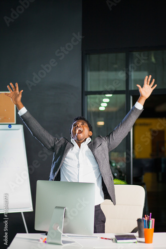 Businessman at office, success gesture, goal reached, happy man. Hardworking male worker. Hipster afro american man at light office with computer monitor. Satisfied with job done