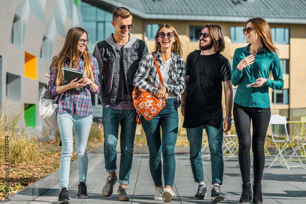 Fototapeta premium Casual style group of students walk along university modern building, have a pleasant talk, sunny day