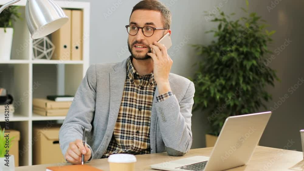 Close up shot of a cheerful man in glasses sitting at the office desk, talking on the phone and holding a pen in his hands. Indoor shot.