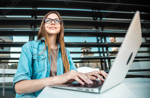 Casual stylish cheerful girl wears jeans scirt works with laptop computer near modern building wall outdoor