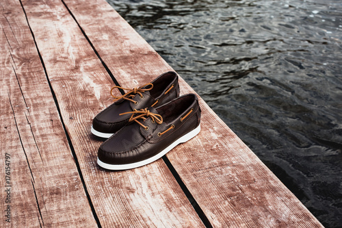 Brown leather men's top sider shoes or boat shoes with white sole on a brown wooden pier or on a brown wooden boards near the water, or rivers, or lakes, or the sea. Fashion advertising shoes photos. photo