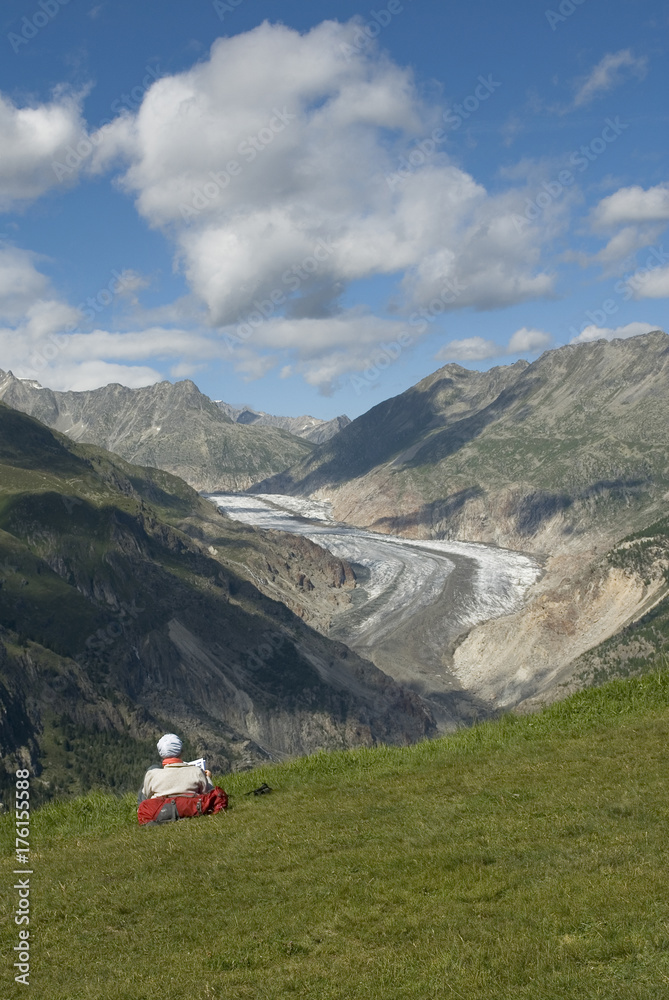 view of Aletsch Glacier, in foreground a middle-aged lady who after a walk reads a book, facing a fantastic landscape, in distance jungfraujoch, eggishorn, eiger, summer, belalp, switzerland.